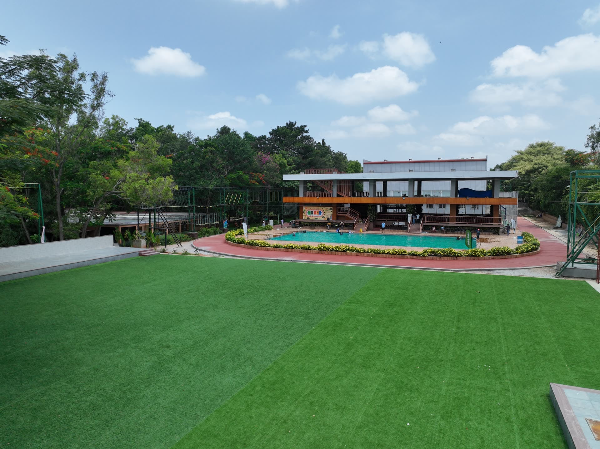 Families arriving on a bright morning at a resort near Electronic City, with games being set up on the lawn beside the swimming pool